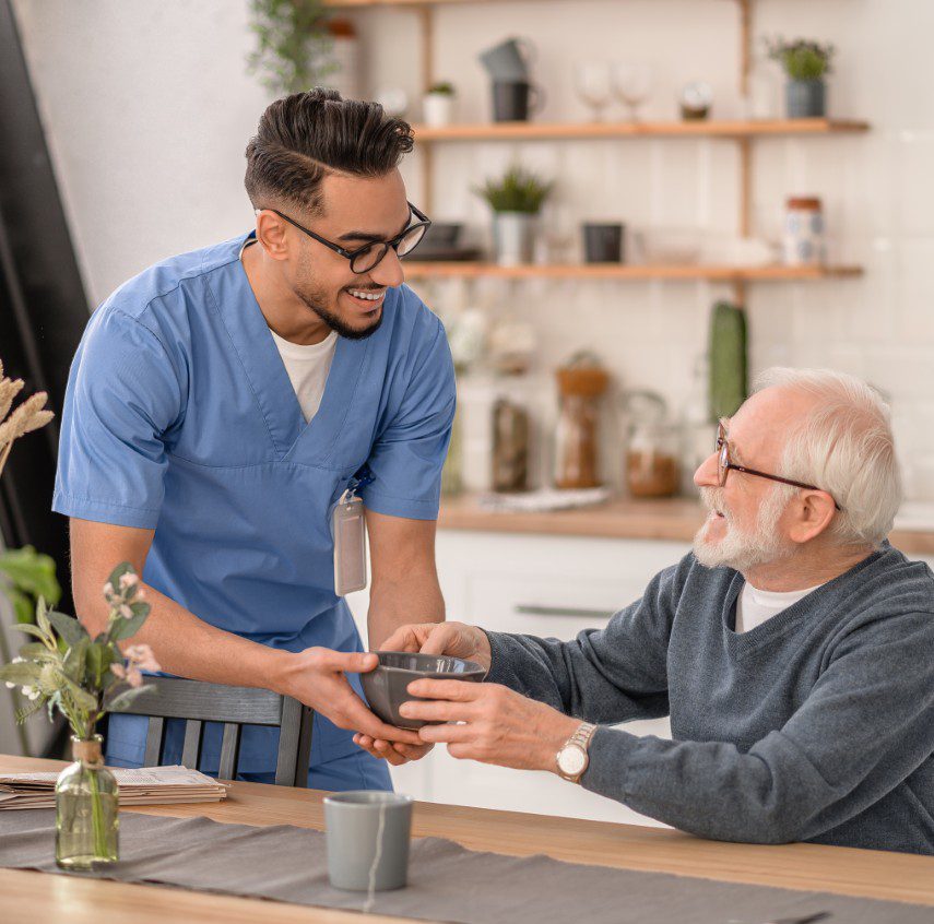 GettyImages-1396691270 Caregiver assisting elderly man in kitchen.